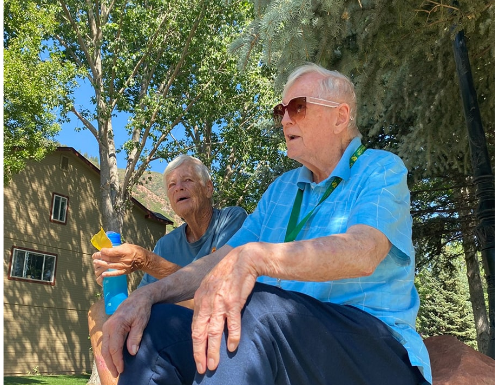 Two elderly men sit outdoors under trees on a sunny day; one wears sunglasses and a blue shirt with a green lanyard, the other holds a blue bottle with a yellow cap.