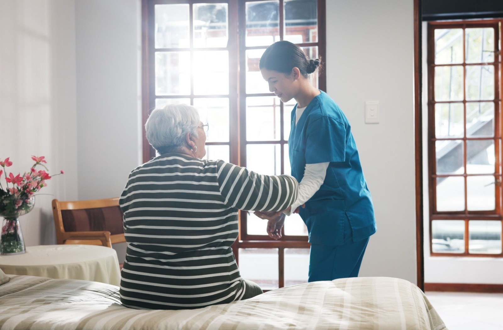 A trained caregiver assists a senior resident with standing up from their bed.
