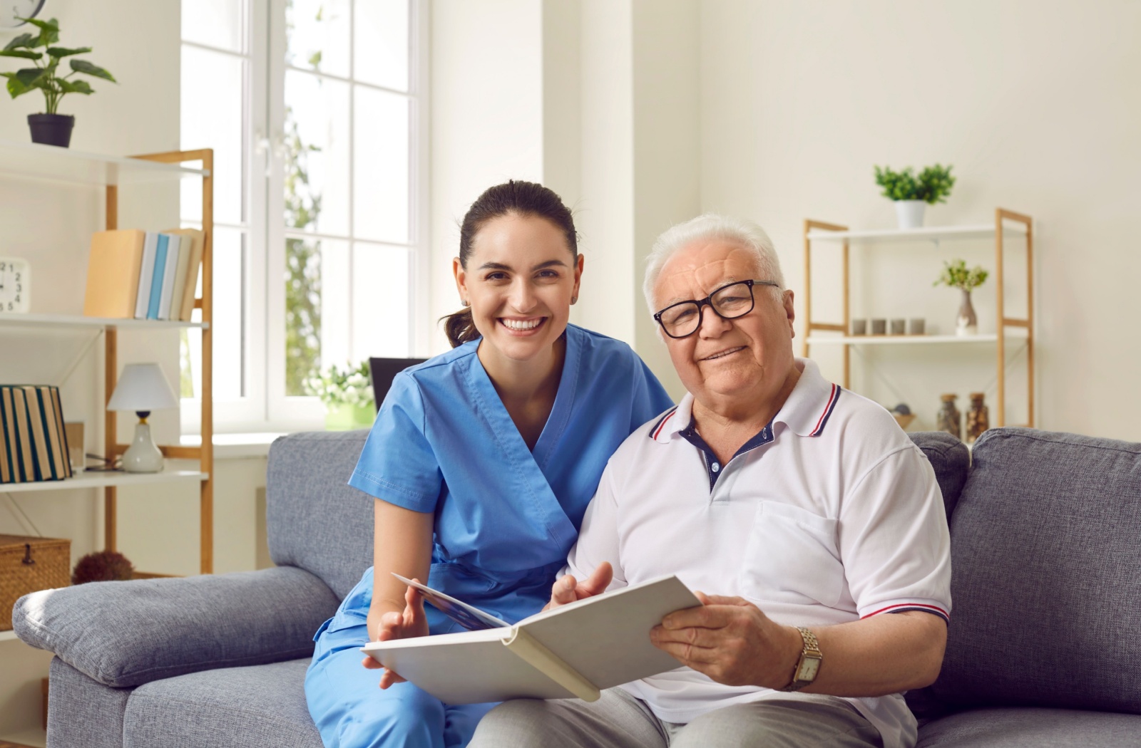 A caregiver and memory care resident smile at the camera while looking through a photo album together.