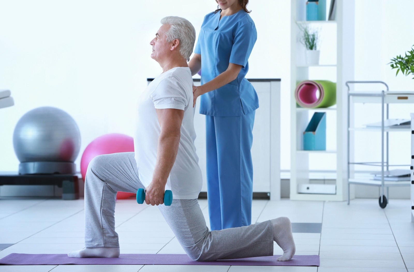 Senior man performing a kneeling lunge with a teal dumbbell while a nurse in blue scrubs supports him in a bright rehab clinic.